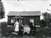Stenhuggare Edvard Nilsson Bohult, Sonen Carl Nilsson, hustru och moder, en släktbild tagen framför ett bostadshus med farstukvist. De bägge äldre personerna sitter vid ett bord på vilket det står ett par kaffekoppar och några vaser.
