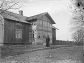 Omålad mangårdsbyggnad med en stor glasveranda i två våningar med snickarglädje längs takfoten.  Framför huset står en kvinna i ytterkläder. Troligen är det vår då lövträden är kala.