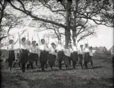 Några kvinnliga gymnaster i blus och s.k. ballongbyxor, som gymnastiserar utomhus. I bakgrunden skymtar några hus. 
Något oskarpt foto.