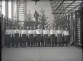 Några kvinnliga gymnaster i blus och s.k. ballongbyxor, uppställda i en stor gymnastiksal. Salen är dekorerad som till fest med granar och
flaggor.