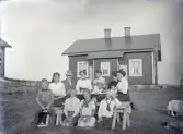 Några sommarklädda barn och vuxna har samlats kring ett enkelt kaffebord utomhus. I bakgrunden syns ett bostadshus, troligtvis hyrt som sommarställe.