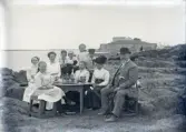 En familj (?) har samlats i det fria, kring ett bord med saft och vatten m.m. I bakgrunden syns Varbergs fästning.