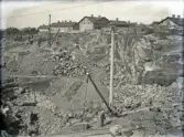 Ett stort stenbrott. I förgrunden syns två män och en lyftkran. I bakgrunden ser man några bostadshus m.m.