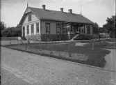 Ett större bostadshus av trä, med snickarglädje och stor veranda. 
I förgrunden syns en nyanlagd trädgård.