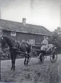 En kvinna sitter med tömmarna i hand i en vagn utanför ett gammalt bostadshus på landet.  Trol. utanför Varberg. 
Suddigt foto.