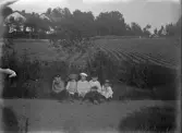Foto av fem barn som slagit sig ner på en smal gräsmatta, utmed en grusgång. 
I bakgrunden syns en åker och några bostadshus.