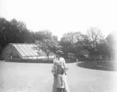 En kvinna och en flicka står på en stor grusplan framför ett växthus m.m.
Troligen vid Ringsberg, Växjö. 
Något otydligt foto.