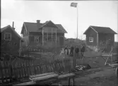 Före detta Södermans gård i Sollenkroka, Vindö. Människor står på gården framför flaggstången med den hissade svenska flaggan. Bilden togs den 10 april 1910.