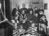 Ett glatt sällskap (familjen Danckwardt med vänner (?), har samlats för gruppfoto i ett hörn av ett vardagsrum (?).
Till vänster skymtar en byrå och till höger syns en kakelugn.
