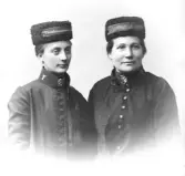 Ateljéfoto av två unga kvinnliga Frälsningssoldater. På vintermössorna sitter ett band med texten 