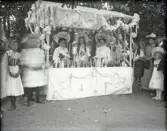 Ett marknadsstånd, dekorerat med girlander m.m. Bakom står tre unga damer i ryschiga stora hattar. 
Framför dem står en mängd vaser med blommor.