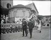 Mycket folk är i rörelse vid Societetshuset. Verandan är prydd med girlander och man skymtar ett stort buffébord. 
Något suddigt foto.