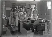 En grupp unga kvinnor iförda stora förkläden och hättor,  i ett skolkök.
Bakom dem syns en svart tavla med recept på¨