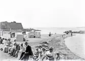 Mycket folk har samlats för att sitta och prata längs strandpromenaden.
I bakgrunden ser man stranden med några badande. Till vänster skymtar Varbergs fästning. 
Något otydligt och suddigt foto.