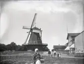 En väderkvarn med ett bostadshus till höger. I förgrunden syns en gammal kvinna och några barn.