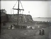 Ett kompani beväringar och några flickor har samlats vid midsommarstången (?) nedanför Varbergs fästning. Kvällsbild.