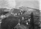 Ett bostadshus (fyrvaktarbostad ?), med fyren som en stor skugga bredvid. Man ser också en del meterologiska mätinstrument, några människor och en segelbåt som ligger förtöjd i bakgrunden.