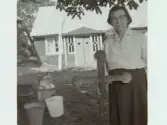 Anna Söder, Friskagården, Friska Bengtsa, Getterön 1170, på Getterön, äldst av gårdarna. Getterön Fjärde vik. Artikel i samband med bilden publicerad i Varbergs Tidning 1958-08-13. I handen håller Anna Söder ett träkors som förr i tiden troddes skydda mot nöd och olyckor. På marken står en mjölkkanna och två spänner.