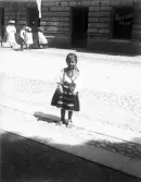 En liten glad flicka, klädd i Rättviksdräkt. Hon står på trottoaren vid en gata. I bakgrunden ser man en kvinna och några andra
flickor.