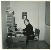 Didners damkonfektion, Växjö.
En kvinna sitter vid ett skrivbord på ett kontor. Hon håller ett par glasögon i handen. I bakgrunden syns en väggalmanacka.