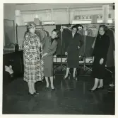 Didners damkonfektion, Växjö.
En kund provar en rutig kappa, assisterad av tre affärsbiträden, varav två främst som smakråd.