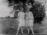 Två unga kvinnor står på ett fält med buskage i bakgrunden i Krogsered. Flickan till höger heter Agnes Johansson och är kusin till fotografen, men kamraten är okänd. Johns faster Augusta och farbror August bodde båda i Krogsered.