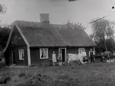 Människor och djur framför stuga med halmtak. Mellan korna står ett äldre par och vid husväggen bakom flickorna står någon sorts motorcykel. Bilden troligen tagen i Gällared eller Okome.