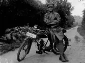Troligen en av bröderna i Breshult poserar med sin motorcykel på en grusväg. Han är klädd för mc-åkning i skinnrock, åtsmitande byxor, kängor och keps.