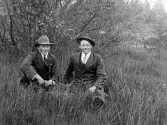 Ynglingarna Gustav Johansson (?) till vänster sitter med sin bror Artur Johansson från Bäckarp, Gällared, i högt gräs ute i naturen.