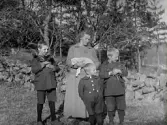 Familjen Johansson i Normanstorp står utomhus i vårsolen, från vänster: Ernst Johansson, mor Amanda Johansson, Georg Johansson, John Johansson. Amanda håller ett nyfött lamm i famnen och två av barnen bär på hönor.