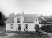 Lantgård med mangårdsbyggnad och bakom den en påbyggd källare. Sticktaken ser ut att vara nylagda. På gräsmattan står späda träd och till höger en motorcykel. Bilden är troligen tagen i Gällared eller Okome.