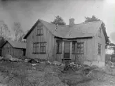 Bostadshus med tresidig farstu i vinkeln. Varken hus eller ekonomibyggnad har målade fasader. Bilden troligen tagen i Gällared eller Okome.