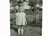 Liten flicka står i en trädgård, troligen Gunhild Andersson från Yngered, Askome, dotter till Teodor och Gerda Andersson.