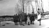 Årnakulla, Krogsered socken. En grupp flickor i olika åldrar står ute i snön utan ytterkläder med händerna i sidorna, framför en kvinna i förkläde. Kanske är det pausgymnastik i en skola? Foto från omkring 1930. Fotografens moster Augusta bodde i Årnakulla.