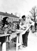 Torghandel i Varberg omkring 1955. Emma Johansson från Träslöv och Nisse Gustavsson. Emma har växter i lerkrukor på bordet och Nisse håller på att så in något i papper.