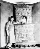 Kakelugnsmakare i arbetskläder framför kakelugn i Tornareds gård, Gällared. I nischen sitter en bebis. Notera kakelugnens fotställning - sådana förbjöds i början av 1800-talet då de ansågs brandfarliga. Kaklet har en snäckrelief, krönet pryds av akantusdekorer och ett manshuvud i centrum medan spiralstavar sitter längs sidorna