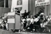 Ordföranden i svenska Unicef-kommittén Lisbeth Palme i talarstolen vid en sammankomst för Unicef i Fässbergsgymnasiets aula i Mölndal, slutet av 1980-talet eller början av 1990-talet.