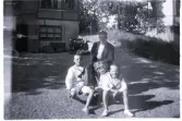 Foto av en man och tre gossar sittandes på gårdsplanen. Text vid bild i album: 