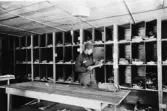 Svenska Frivilligkåren i Finland.  Fältpostkontor i Tornio,
1940.  Brevavdelningen.  Korsbandssorteraren.  Korsbandsfacken av
masonite och omställbara.  Bl.a. fördelades c:a 500 oadresserade
tidningar per dag.  Stående: Postbiträdet Isaksson fr. Dorotea.