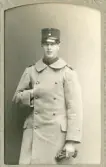 En man i uniform (m/23?) med uniformskappa och kepi.
Knäbild, en face. Ateljéfoto.