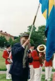 Nationaldagsfirande i Stadshusparken i Mölndal den 6 juni 1999. Lars Gahrn bär den svenska flaggan. I bakgrunden Mölndals Paradorkester.
