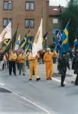 Nationaldagsfirande i Stadshusparken i Mölndal den 6 juni 1999. Här ses en samling människor tåga in på Häradsgatan med svenska flaggor och föreningsflaggor.