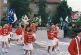 Nationaldagsfirande i Stadshusparken i Mölndal den 6 juni 1999. Här ses en samling människor tåga in på Häradsgatan. I förgrunden Mölndals Paradorkester, i bakgrunden svenska flaggor och föreningsflaggor.