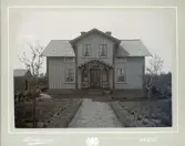 Ett bostadshus med veranda i en trädgård. Till vänster syns ett uthus. 
Fastigheten Bergslund 1, Berg.