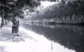 Foto av en kvinna som står vid en kajkant med kanalbåtar i bakgrunden, troligen i Amsterdam, 1924-25.