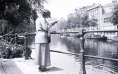 Foto av en kvinna som står och läser en bok vid en kajkant, troligen i Amsterdam, 1924-25.