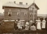 Foto av prästgården i Falkenberg c:a 1902  Sittande fr. v.: kyrkoherde D Davidsson hans dotter Maria, fru Davidsson f. Lundgren, fröken Skogman (tillfälligt där för att lära hushåll) gamla fru Davidsson dvs. Sara Arvidsdotter, kyrkoherdens mor.  Stående bakom henne Anna Davidsson, gift Hallbäck, två tjänarinnor okända, på gräsmattan sitter Axel o Edit Davidsson, brorsbarn till kyrkoherden och de kallades fosterbarnen. Axel dog ung, Edit blev lärarinna i Norrland, Clara Hellgren, bodde i prästgården. (A.S.)  På baksidan av fotot står: 3544 f) Gåva av W E Erlandsson, Kvidinge sittande fr.v. 1 Kyrkoherde David Davidsson 4. Gerda Skogström ( vit blus).