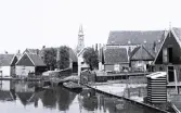 Foto av en stadsmiljö med en kanal i förgrunden, hus längs vattnet och en kyrka med ett spetsigt torn i bakgrunden, någonstans i Holland, 1924-25.