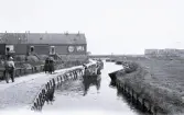 Foto av de traditionella nederländska fiskebyarna där kanalen kantas av byggnader och en gångväg där människor syns. I vattnet ligger det små båtar och två kvinnor för fram en båt, 1924-25.