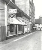 Tiden är 1934. (textkompletering E Kylén).  På affärsskyltarna kan man läsa, Allan Johansson Skomakare, Carl Ander Tapetserar Verkstad, Almqvist Foto, Frisör, Elktrotekniska byrån Walter Bengtsson.